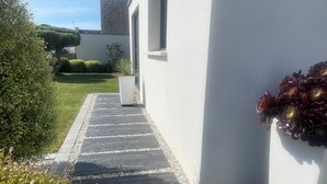 Un sentiero conduce a un edificio con facciata bianca, una pianta in vaso e un giardino con arbusti e fiori.