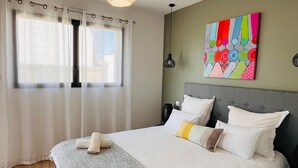Un letto rifatto con lenzuola bianche, una coperta grigia e due asciugamani arrotolati. Sopra il letto, un quadro astratto dai colori vivaci.