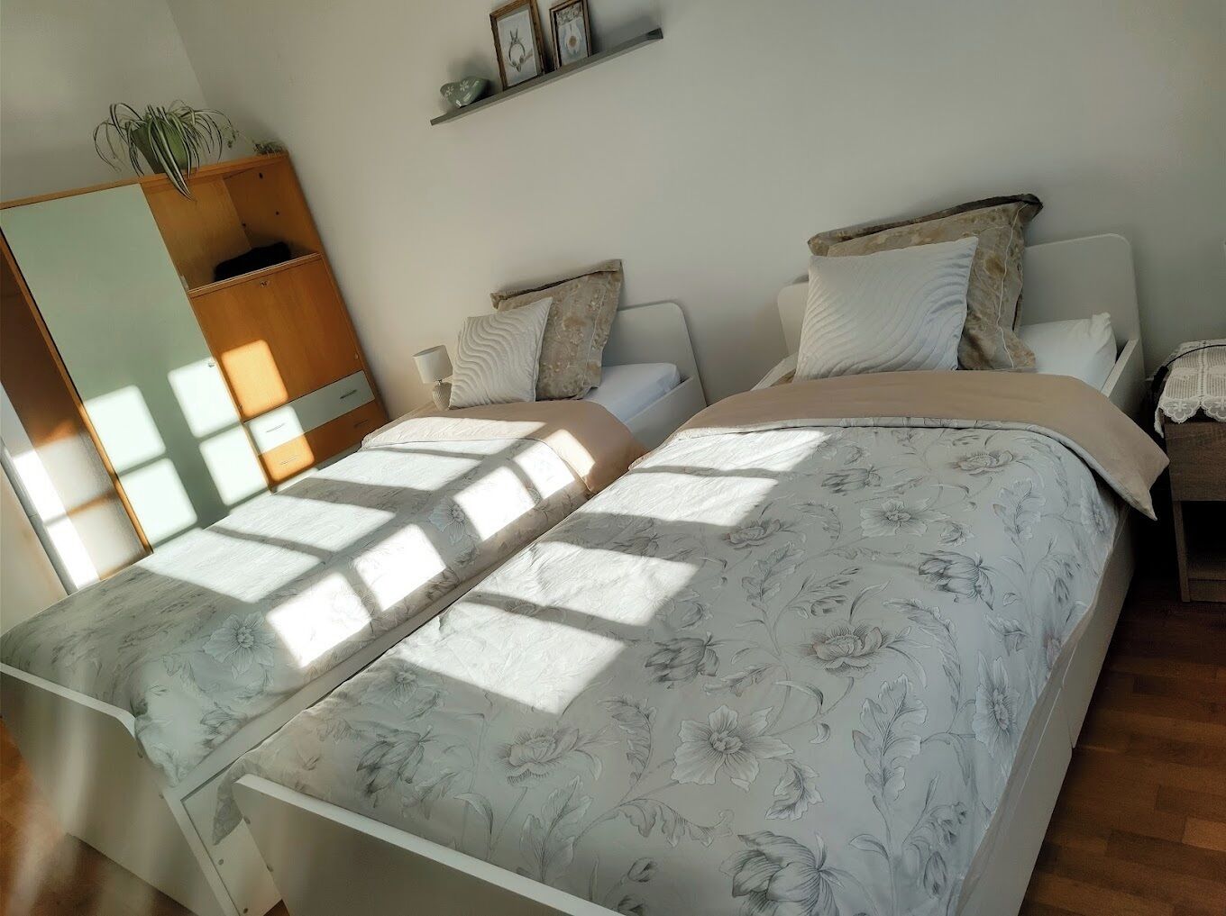 A bedroom with two beds, a wooden wardrobe, a bedside table, and a shelf with pictures.
