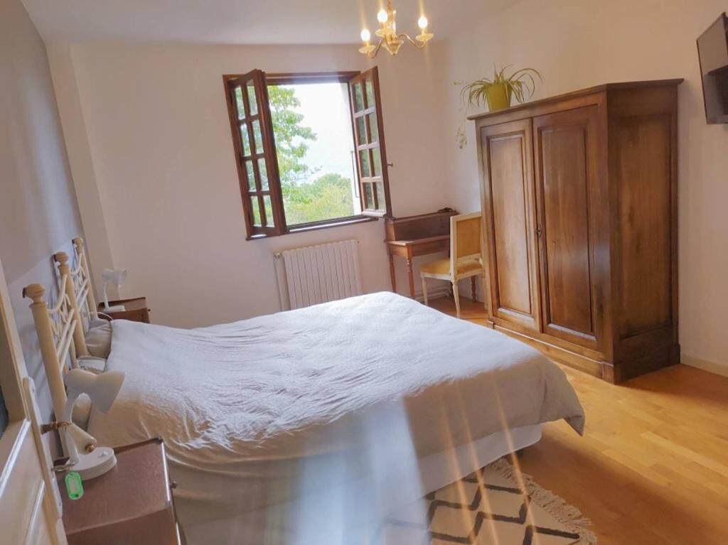 Un dormitorio con cama, armario de madera, radiador, ventana con cortinas y una pequeña mesa con silla.