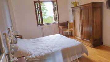 Un dormitorio con cama, armario de madera, radiador, ventana con cortinas y una pequeña mesa con silla.