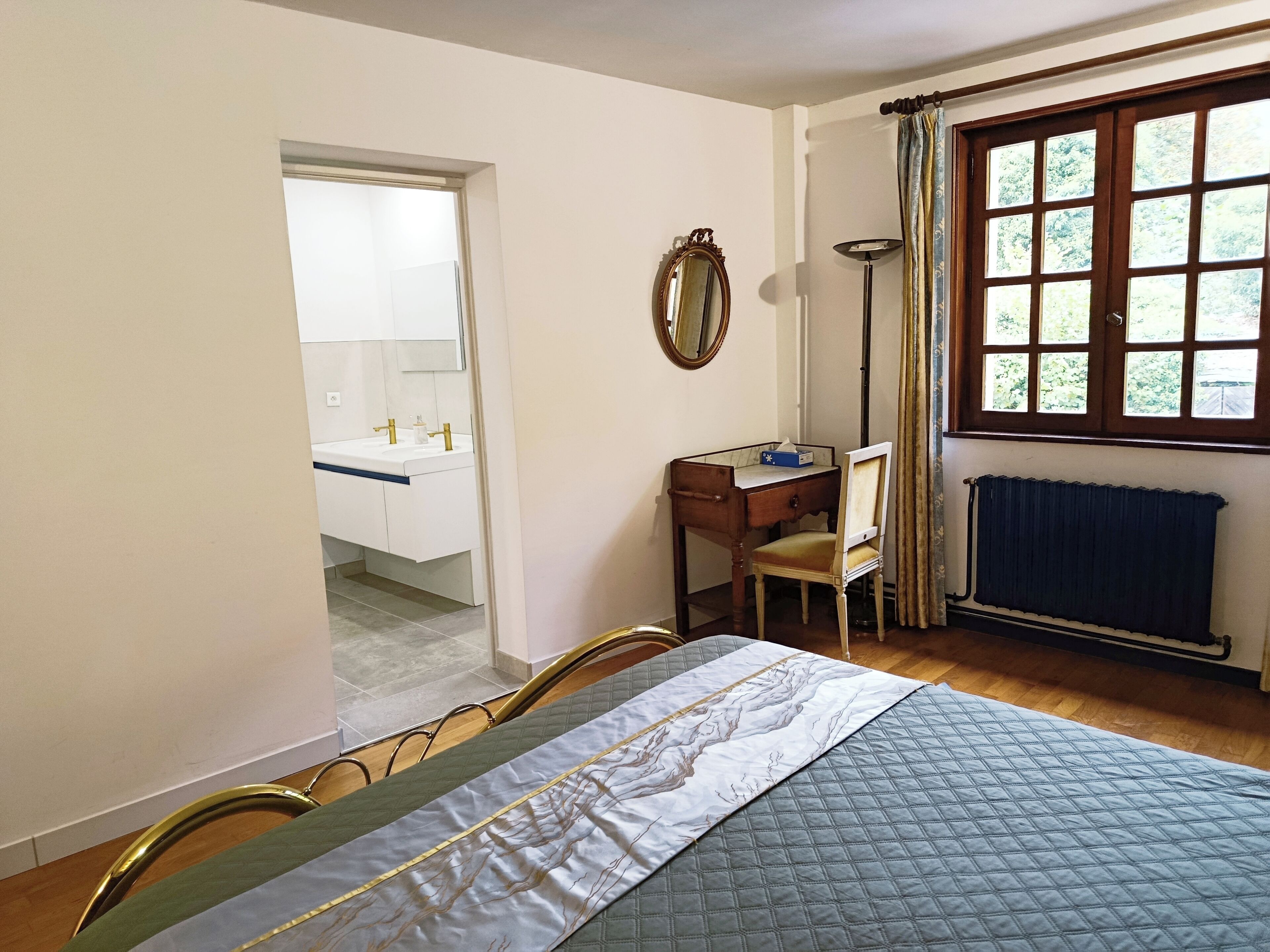 A bedroom with a bed, a desk, a chair, a mirror, a window with curtains, and a radiator.