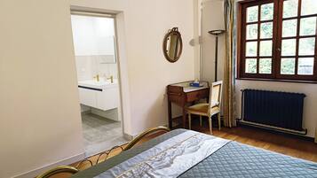 A bedroom with a bed, a desk, a chair, a mirror, a window with curtains, and a radiator.