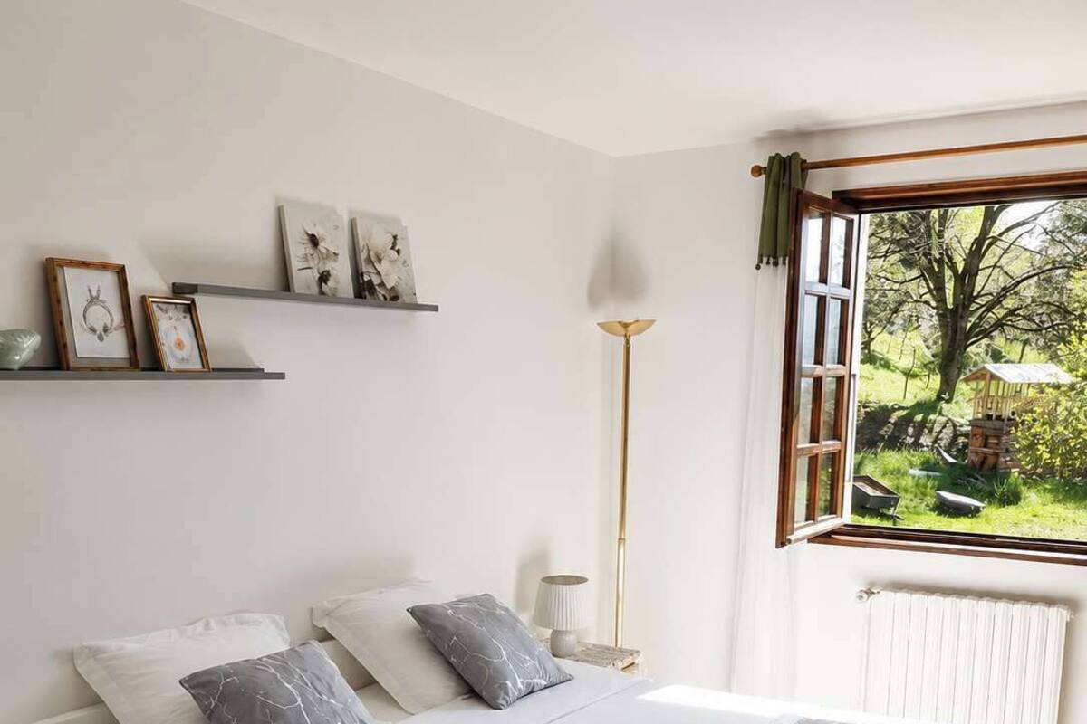 A bedroom with a bed, bedside tables, a floor lamp, a window with a view of greenery, and wall-mounted shelves with framed pictures.
