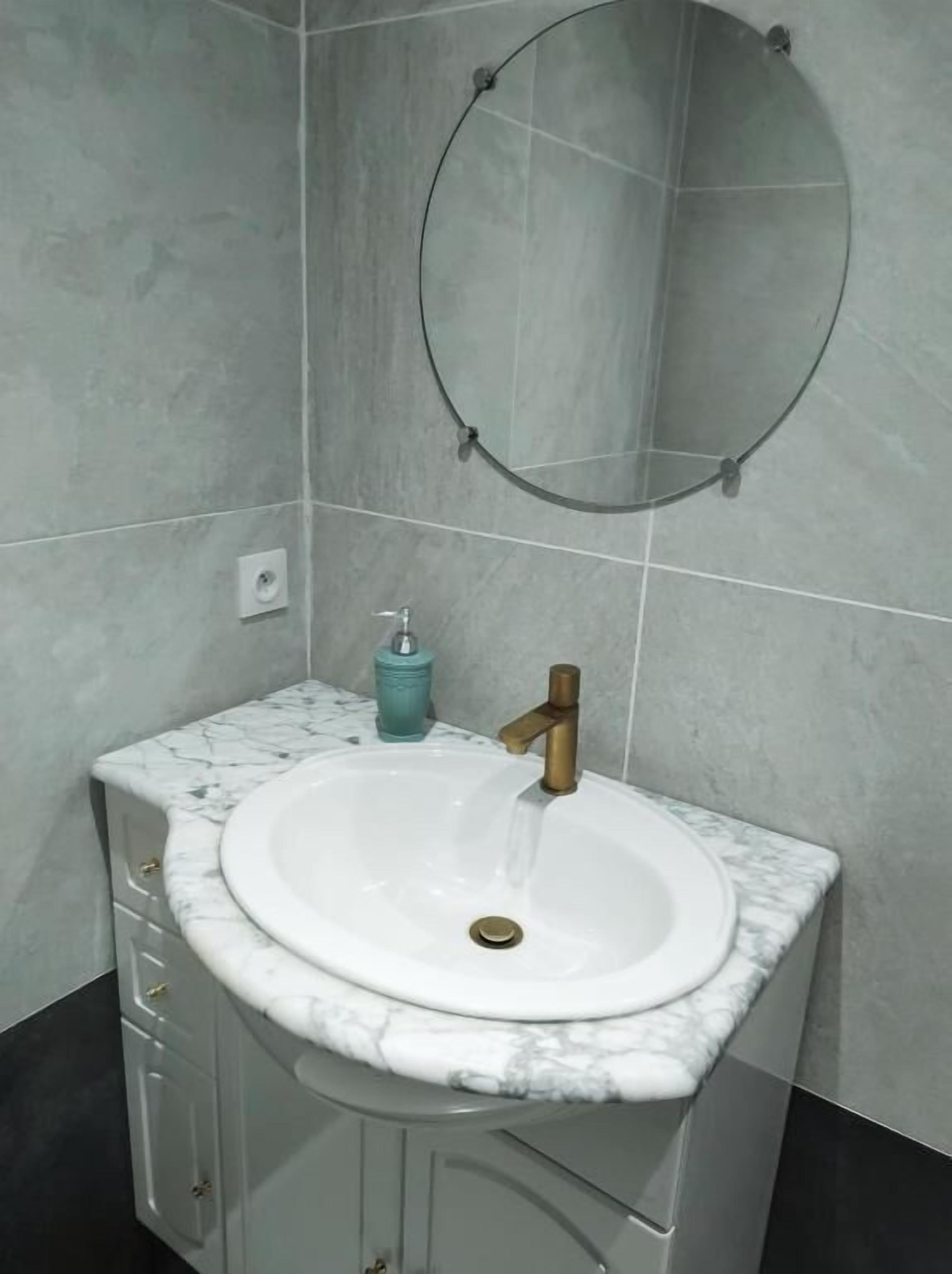 Un baño con lavamanos de mármol, grifo de bronce, espejo redondo y dispensador de jabón color aguamarina.