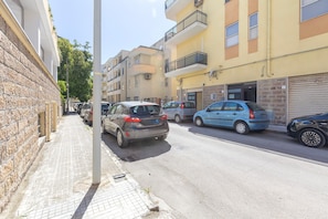 Eine schmale Stadtstraße gesäumt von mehrstöckigen Gebäuden, parkenden Autos und einem Bürgersteig.