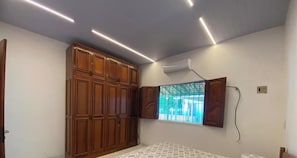 A bedroom with a large wardrobe, an air conditioning unit, a window with bars, and a bed with a patterned bedspread. - Casa de Praia Na Ilha de Marajó - Joanes/PA (Salvaterra)