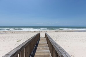 Ein hölzerner Steg führt zu einem Sandstrand, im Hintergrund das Meer.
