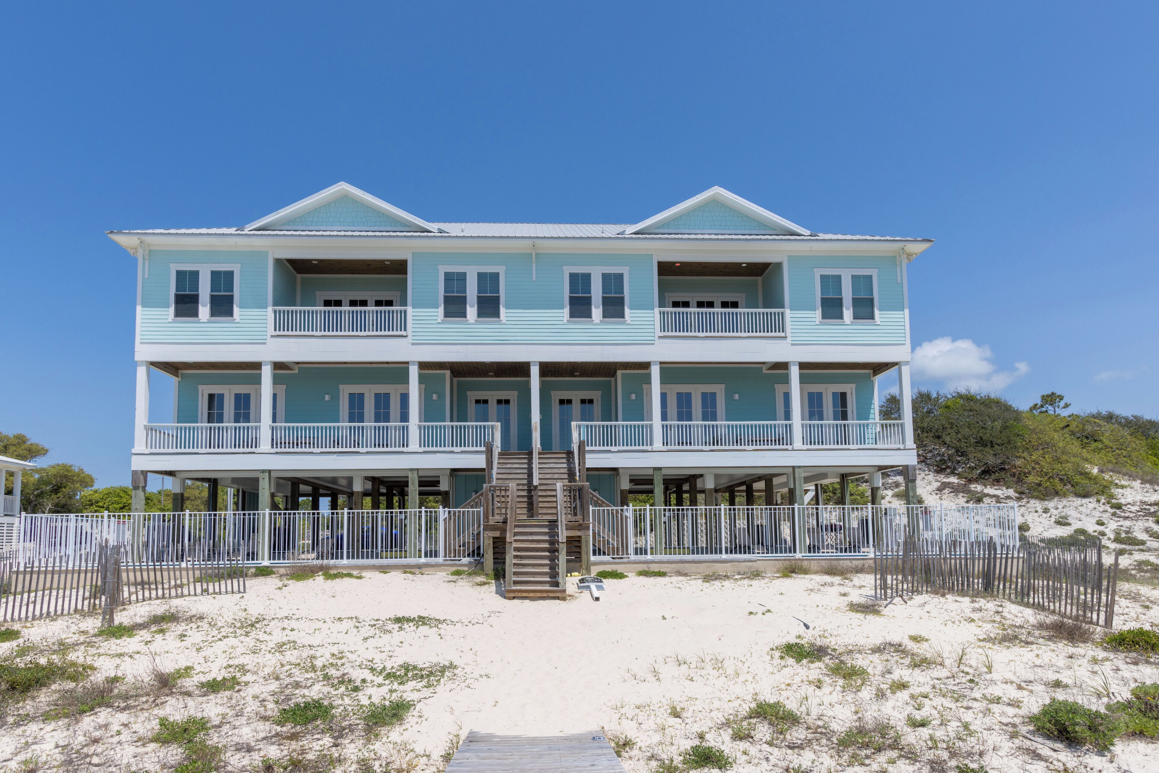 Ein zweistöckiges Strandhaus mit einer zentralen Treppe, mehreren Balkonen und einem Sandstrand im Vordergrund.