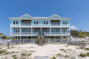 Ein zweistöckiges Strandhaus mit einer zentralen Treppe, mehreren Balkonen und einem Sandstrand im Vordergrund.