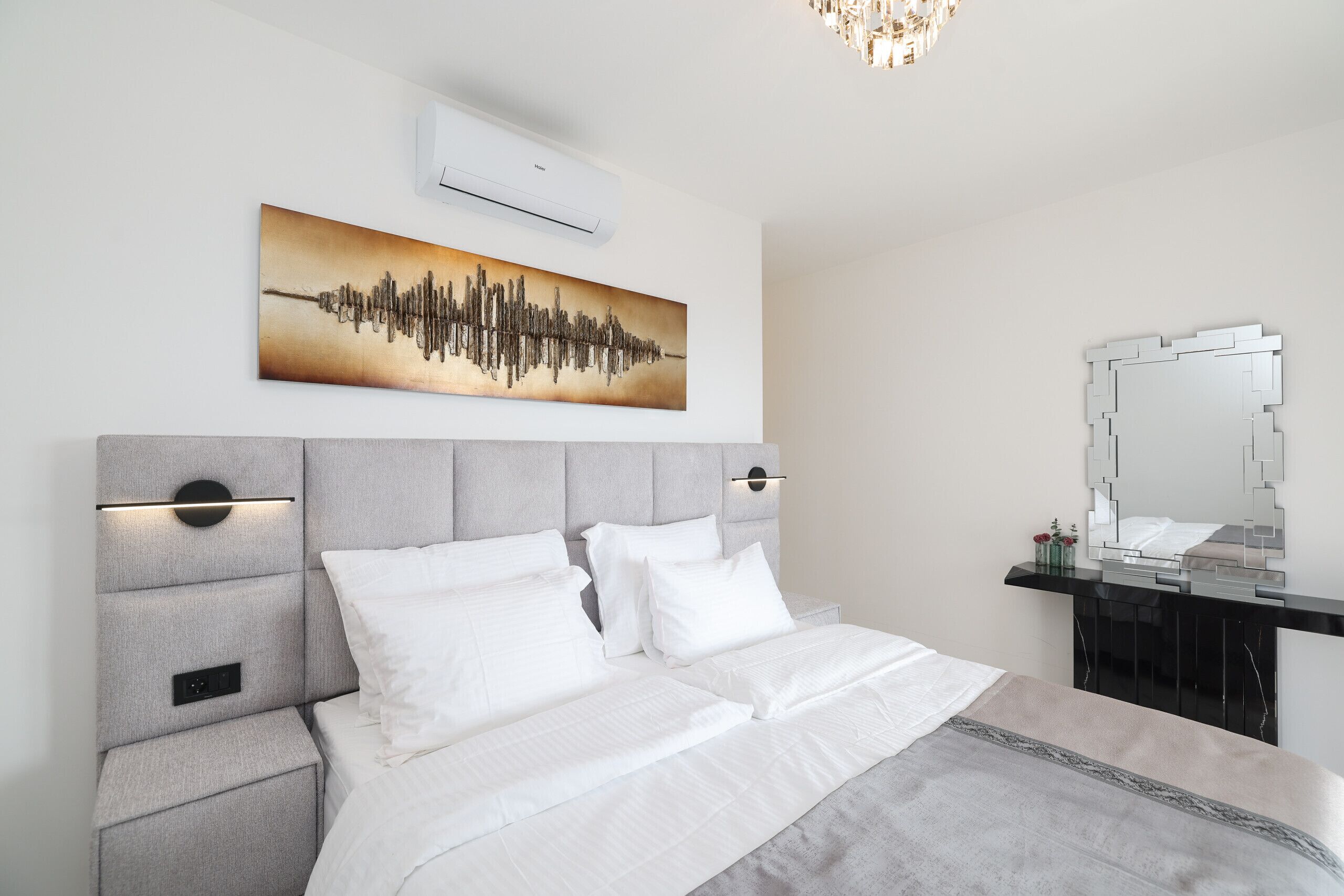 Un dormitorio moderno con cama, mesitas de noche, aire acondicionado de pared, un cuadro abstracto grande y un espejo sobre un tocador.