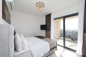 Un dormitorio con cama, televisor, candelabro, balcón con una silla y vista a la ciudad.