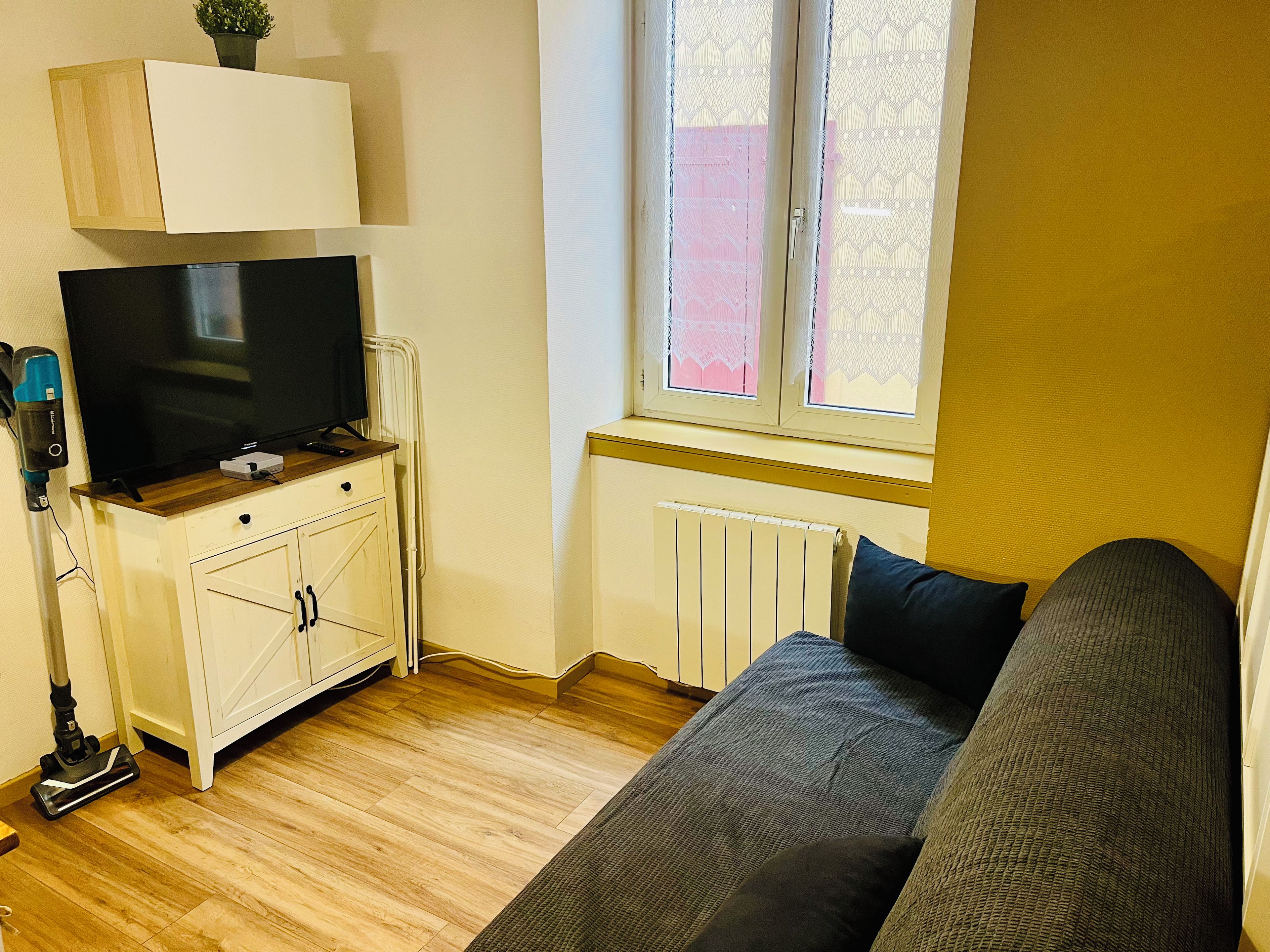 Una habitación pequeña y funcional con televisión sobre un mueble de madera, un radiador, una ventana con cortinas de encaje y un sofá.