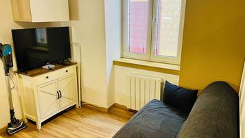Una habitación pequeña y funcional con televisión sobre un mueble de madera, un radiador, una ventana con cortinas de encaje y un sofá.