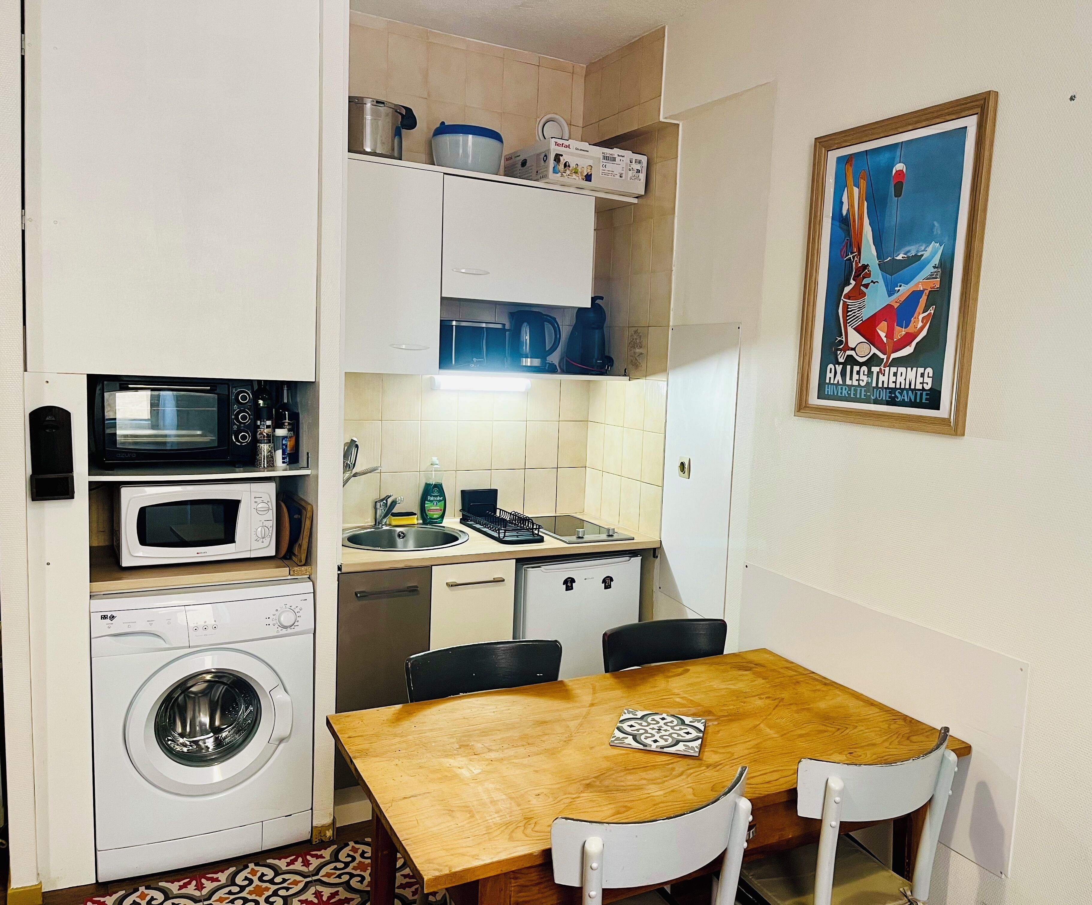 A compact kitchen with a washing machine, two microwaves, a sink, and a dining table with chairs.