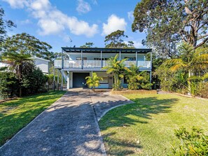 A two-story house with a balcony, surrounded by lush greenery and a paved driveway.