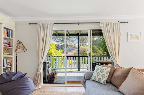 A room with a sofa, a bookshelf, a window with curtains, and a view of a balcony with outdoor furniture.