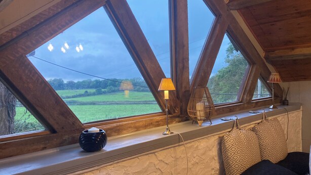 A room with a sloped ceiling, a window with a view of a green field, a lamp, a decorative item, and patterned pillows. - The Stable, Sleeps 2 With Parking (Llanfairpwllgwyngyll)