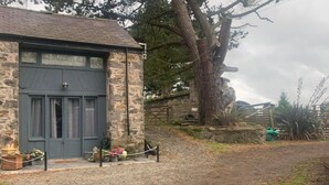 A stone building with a grey door and windows, a paved driveway, and a large tree in the background. - The Stable, Sleeps 2 With Parking (Llanfairpwllgwyngyll)