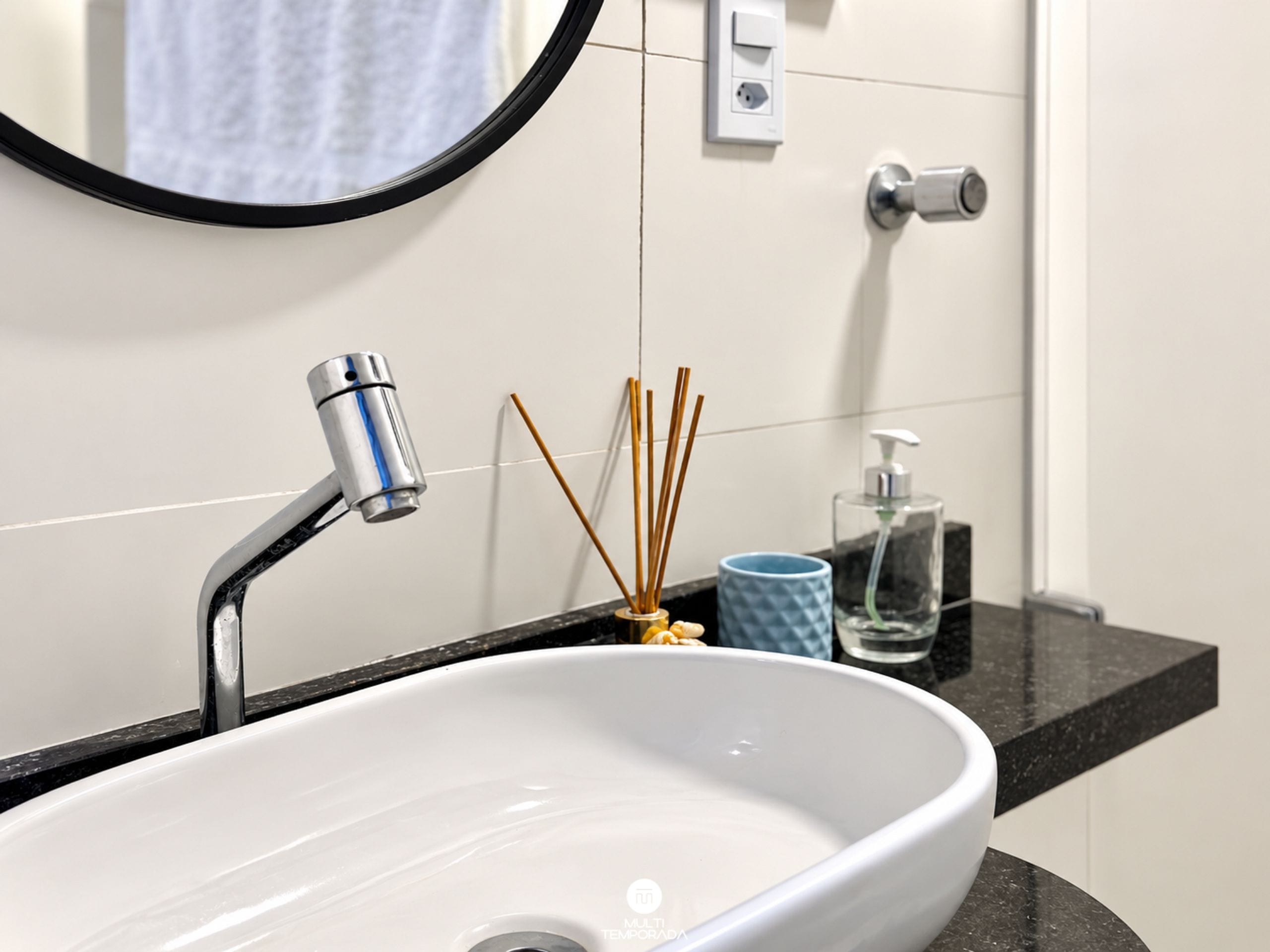 Een badkamer met een witte wasbak, een ronde spiegel, een moderne kraan, een zeepdispenser en een blauw kopje.