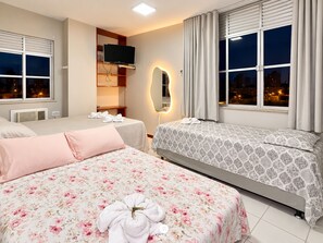 Habitación de hotel con dos camas, un televisor montado en un estante de madera, un espejo y vistas a la ciudad desde la ventana.