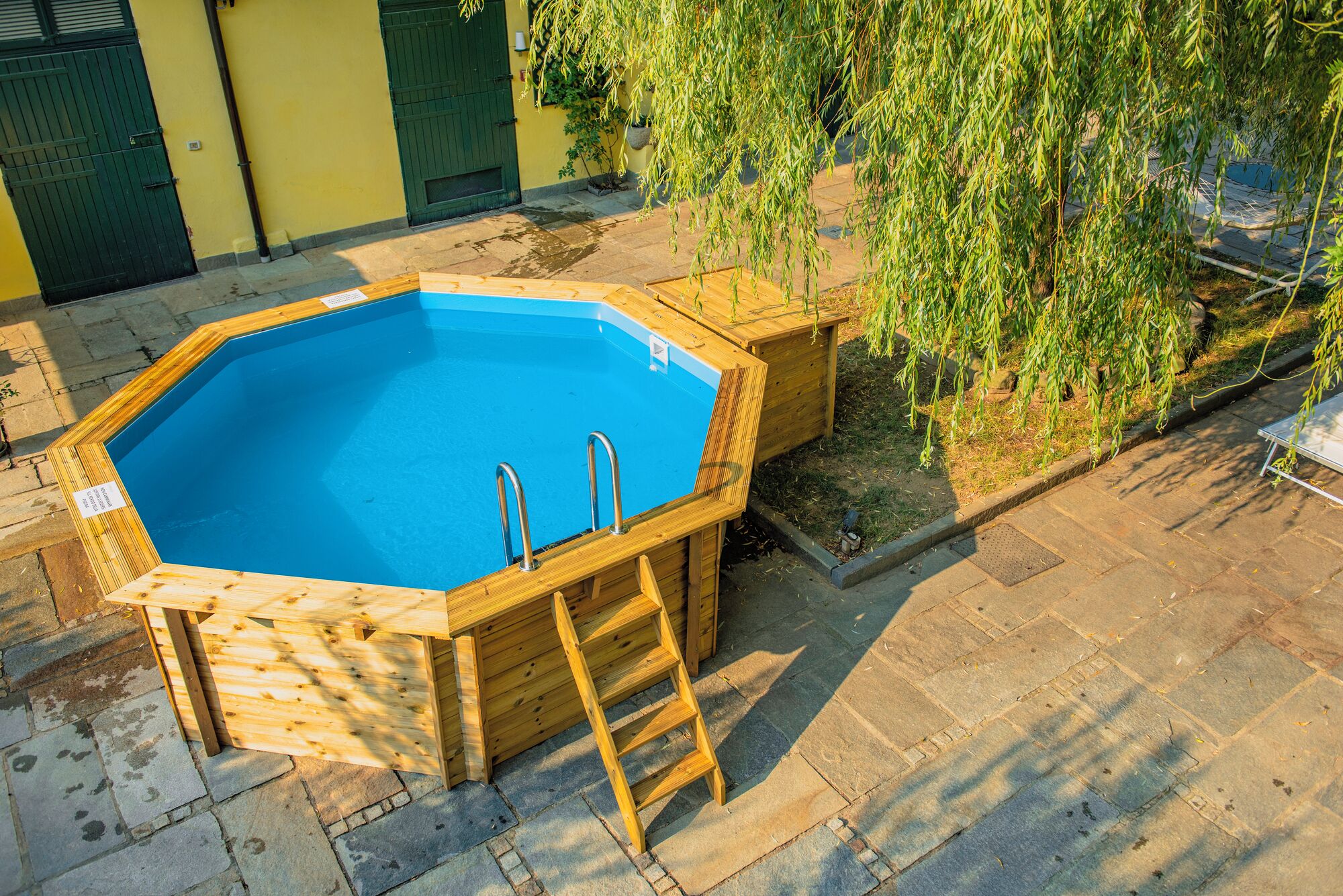 Una piscina hexagonal de madera con escalera, rodeada de baldosas de piedra y una puerta verde.
