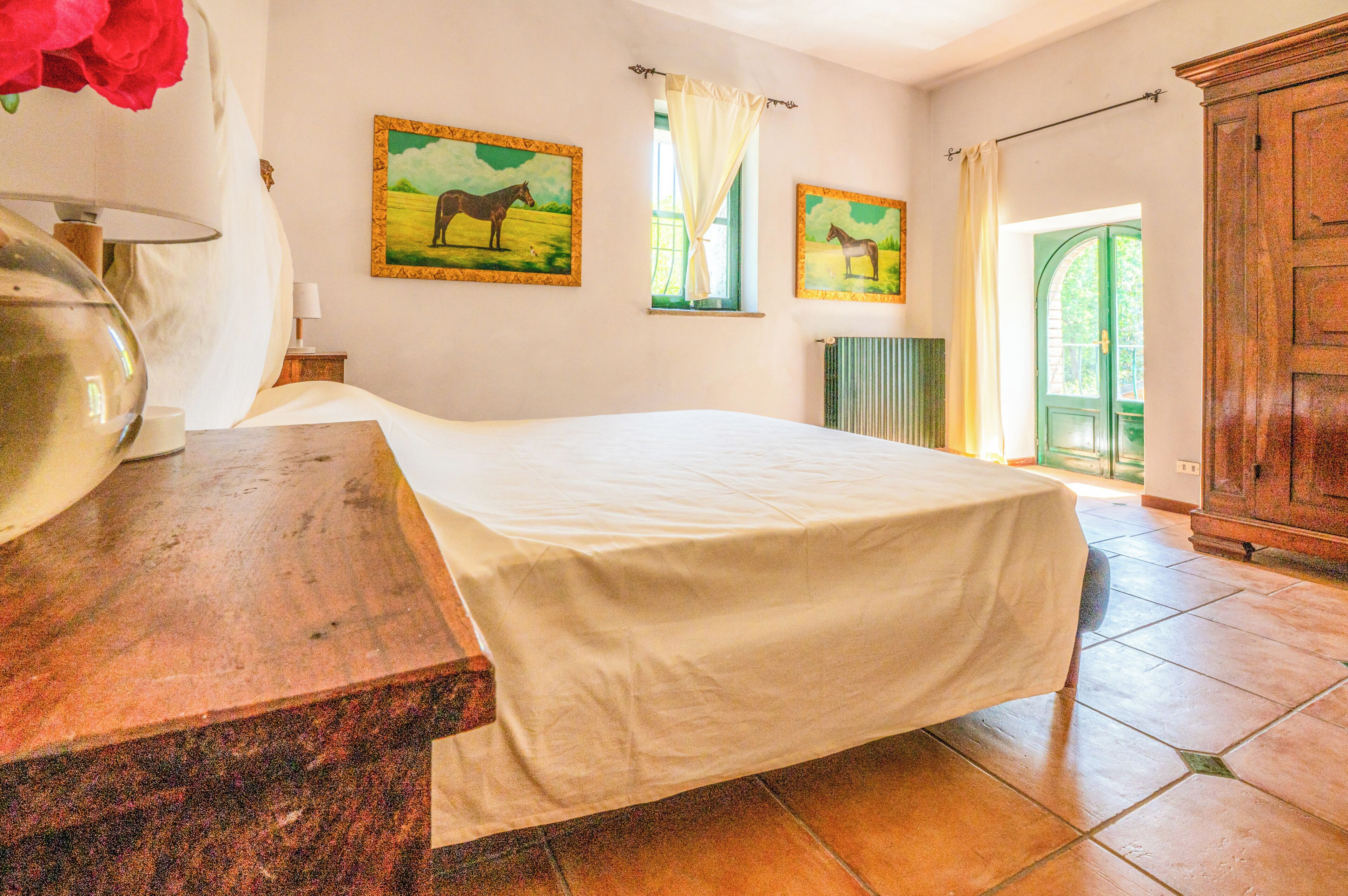 Une chambre à coucher avec un lit, une table de chevet en bois, un vase avec des fleurs, deux tableaux représentant des chevaux, une fenêtre avec des rideaux et un radiateur.