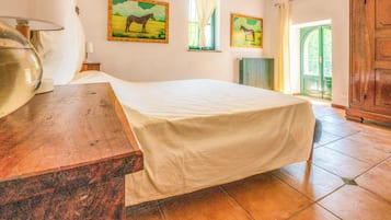 Une chambre à coucher avec un lit, une table de chevet en bois, un vase avec des fleurs, deux tableaux représentant des chevaux, une fenêtre avec des rideaux et un radiateur.