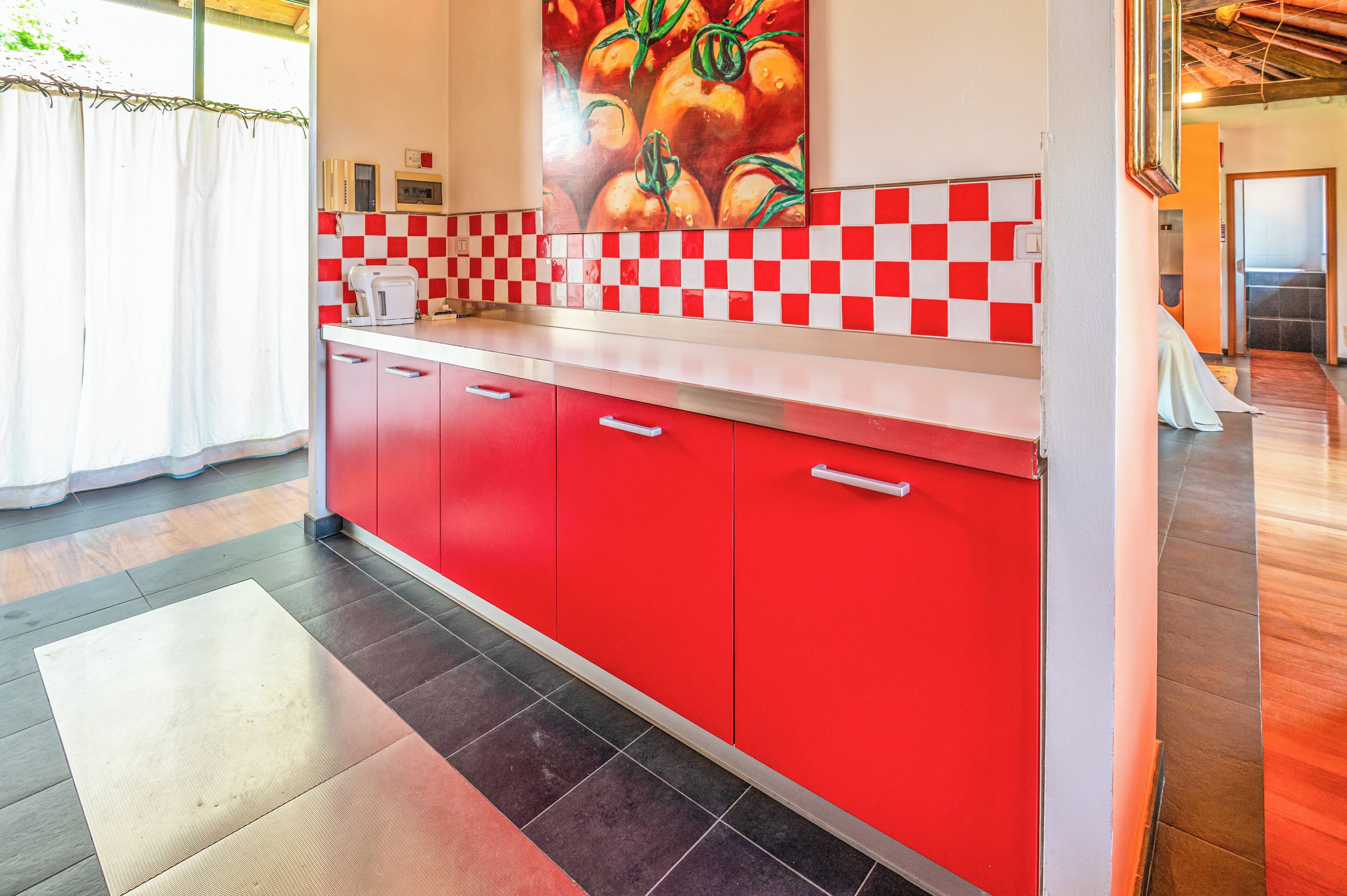 A kitchen with red cabinets, a checkered backsplash, and a painting of tomatoes.