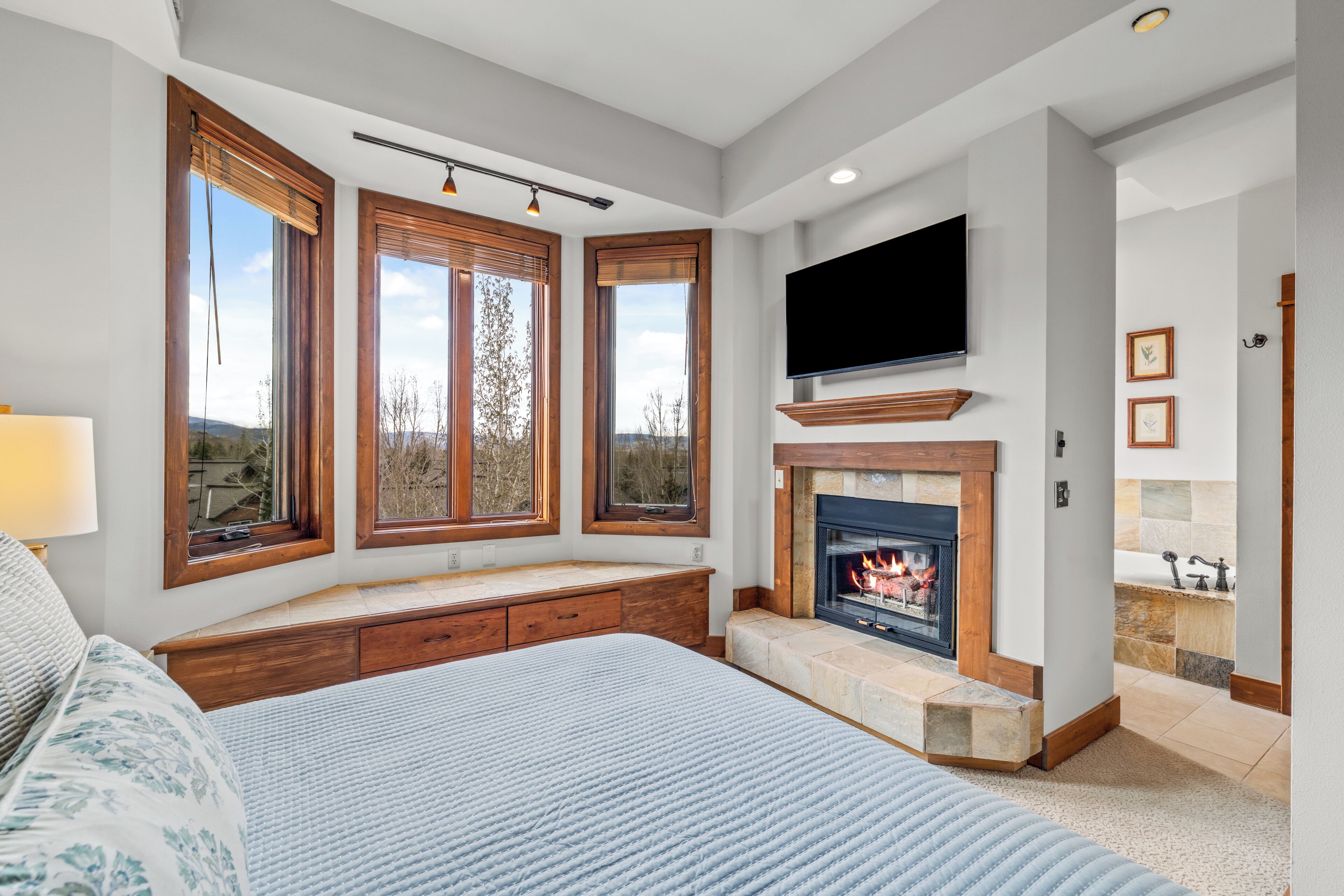 A bedroom with a large bed, a fireplace, a TV, and a window with a view.