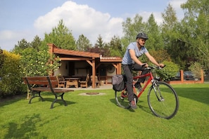 A person riding a bicycle in a grassy area with a wooden structure and a bench in the background.