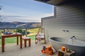Un balcón con mesa de madera, una silla de mimbre, una bañera grande y vistas a colinas ondulantes.