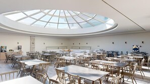 Un comedor amplio con varias mesas y sillas, un tragaluz central y una cocina moderna a la vista.