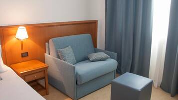 A hotel room with a single blue sofa, a wooden nightstand with a lamp, a small table, and a grey ottoman.