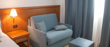 A hotel room with a single blue sofa, a wooden nightstand with a lamp, a small table, and a grey ottoman.