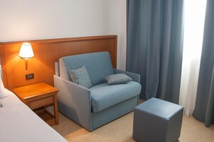 Habitación de hotel con un sofá azul individual, una mesita de noche de madera con una lámpara, una pequeña mesa y un otoman gris.