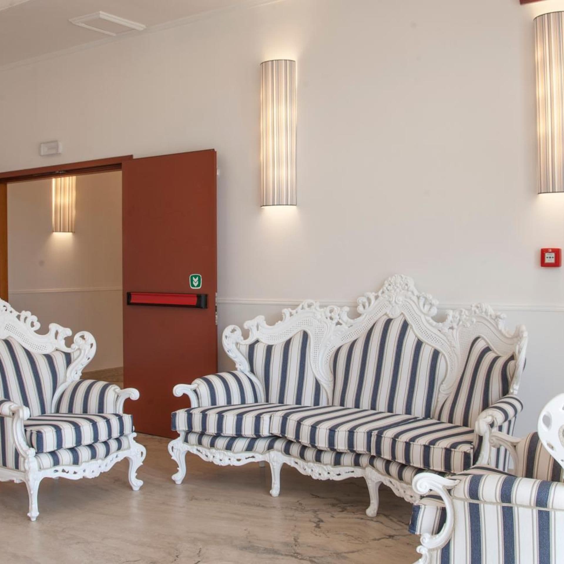 A seating area with striped upholstered chairs and sofas, a wooden door, and wall-mounted light fixtures.