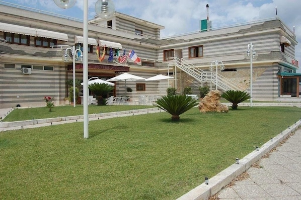A hotel with a flat roof, multiple floors, and a central staircase. There are outdoor seating areas with umbrellas.