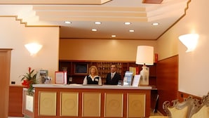 Zona de recepción de hotel con mostrador, dos empleados, escritorio de recepción, lámpara y área de descanso con sofá.