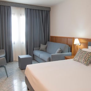 Una habitación de hotel con una cama, un sofá, una silla, una mesita de noche y una lámpara.