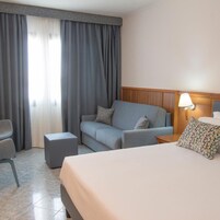 Una habitación de hotel con una cama, un sofá, una silla, una mesita de noche y una lámpara.