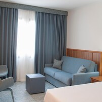 Una habitación de hotel con un sofá gris, un pequeño reposapiés, una mesita de noche de madera y una cama.