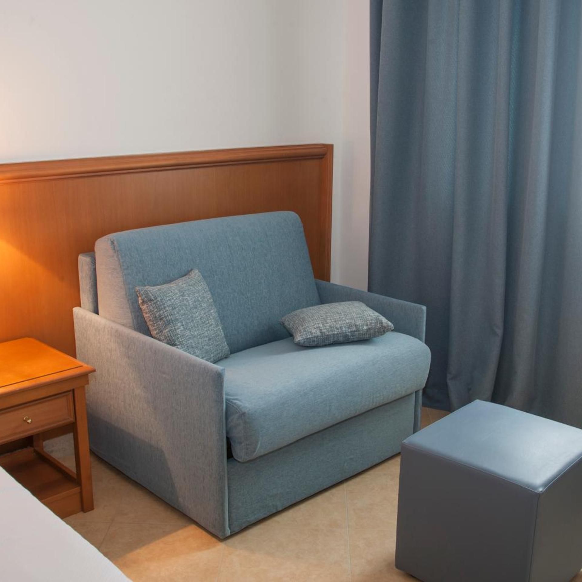 A room with a blue sofa, a wooden nightstand, a small ottoman, and a curtain.