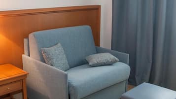 A room with a blue sofa, a wooden nightstand, a small ottoman, and a curtain.