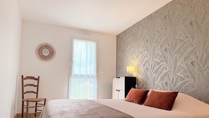 A bedroom with a bed, a chair, a wall-mounted lamp, and a patterned wall. - The Liberators - Collection La 21ème Planche (Bayeux)