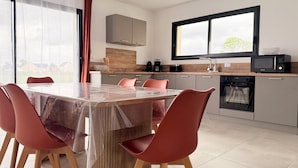 A modern kitchen with a dining area, featuring a large table, red chairs, a microwave, and an oven. - The Liberators - Collection La 21ème Planche (Bayeux)