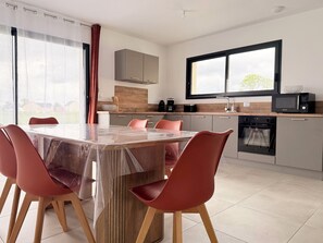 A modern kitchen with a dining area, featuring a large table, red chairs, a microwave, and an oven. - The Liberators - Collection La 21ème Planche (Bayeux)