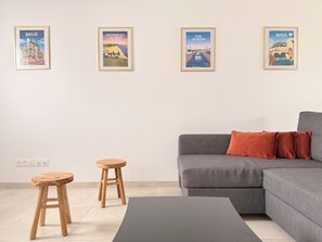 A modern living room with a grey sofa, two wooden stools, a black coffee table, and four framed posters on the wall. - The Liberators - Collection La 21ème Planche (Bayeux)