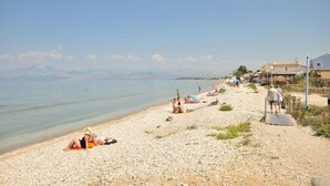A pebbled beach with people sunbathing and walking, a clear sky, and distant mountains. - Elena Apartments (Corfu)