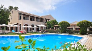 A swimming pool with lounge chairs and umbrellas, surrounded by a building with balconies and a garden. - Elena Apartments (Corfu)